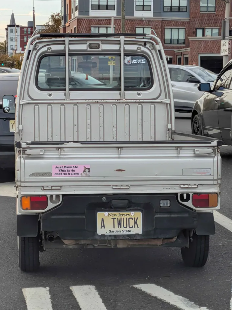 a kei truck with a NJ license plate that says A TWUCK and a bumper sticker that says Just Pass me This is as Fast as it Gets